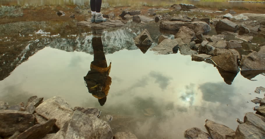 Young female photographer with a backpack and camera standing on rugged rocks by a tranquil mountain lake, taking in the stunning landscape under a cloudy sky