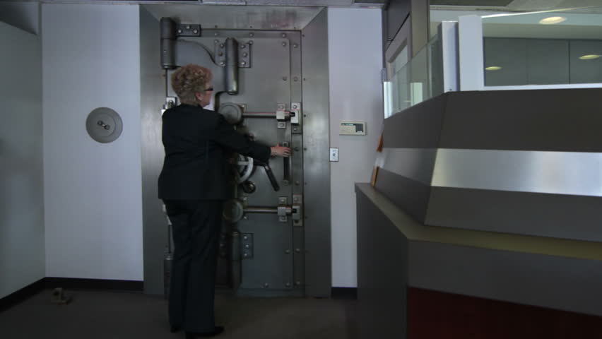 Woman opens a large bank vault door revealing the safe deposit boxes inside.