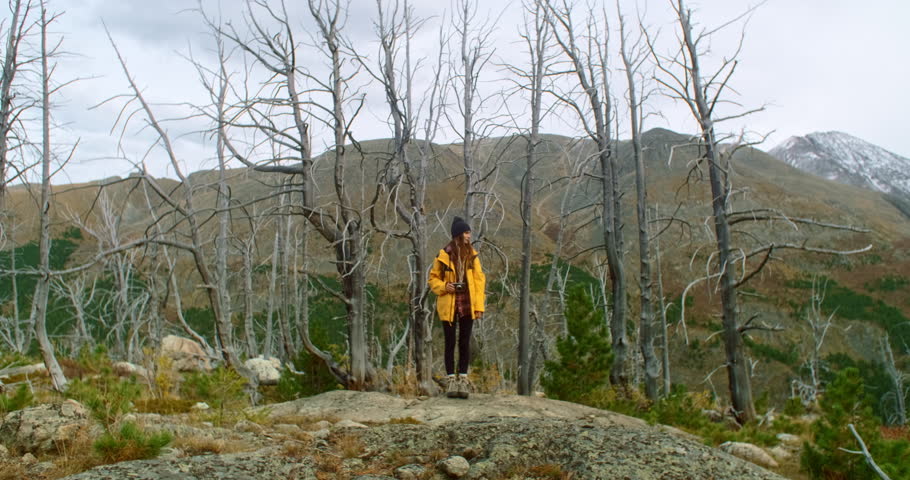 Photographer wearing a yellow jacket, beanie and boots is standing on a rock, holding a camera and taking pictures of a mountain landscape with dead trees