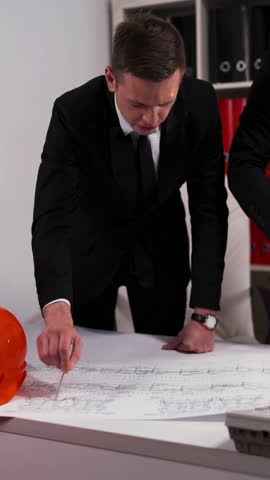 Two handsome attractive engineers businessmen in business office look at architect drawing with helmet they discuss about new construction. 4k UHD Vertical Shot.