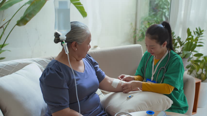 Asian young nurse putting bandage on senior patients arm while giving her intravenous vitamin course infusion during home visit