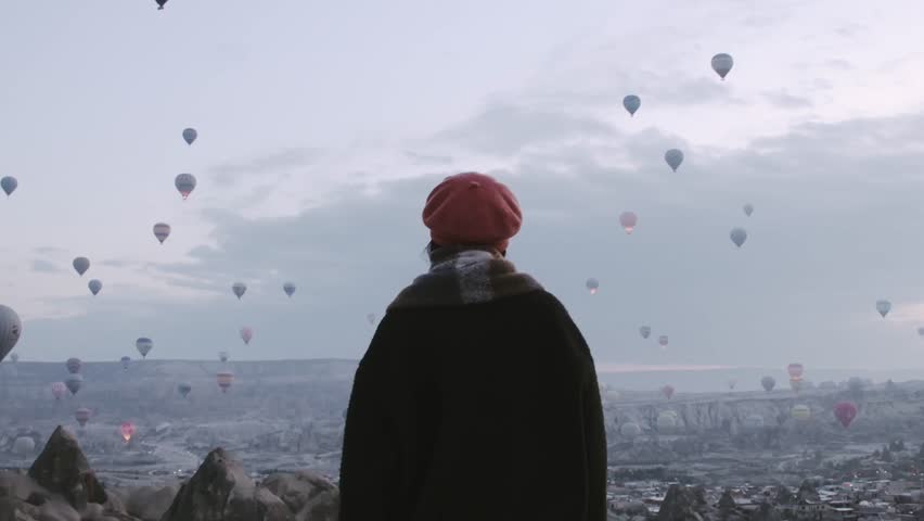 Close up back view slow motion Woman looking around observe,wear fashionable clothing stand on scenic viewpoint watch hot air balloon flight on sunrise with beautiful goreme town and valley background