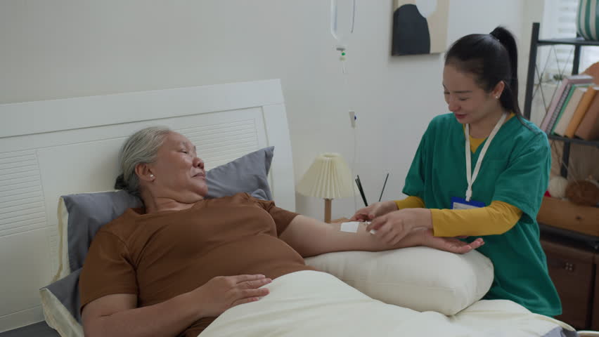 Young female physician helping Asian senior lady in receiving IV drip infusion and vitamin therapy while providing home service