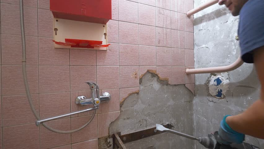 Construction workers remove old pink tiles from bathroom wall using electric hammer. Process showcases renovation and demolition techniques in home improvement projects