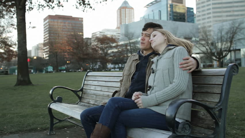 Attractive young couple sitting on a bench, talking, smiling, embracing with the