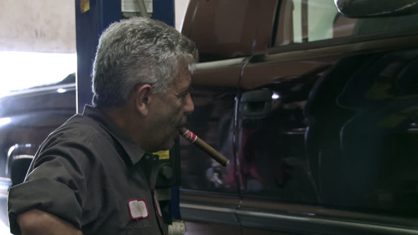 Auto mechanic smokes a cigar as he lowers a car on a hoist.