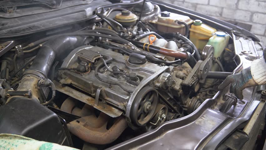 Car mechanic inspects steaming engine compartment while wearing gloves. Using tool, mechanic checks for issues in engine, addressing potential overheating problems