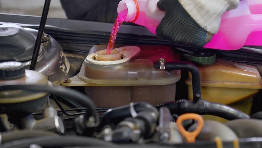 Mechanic wearing gloves pours pink coolant into car engine, ensuring proper maintenance and preventing overheating. Process involves filling reservoir with essential fluid