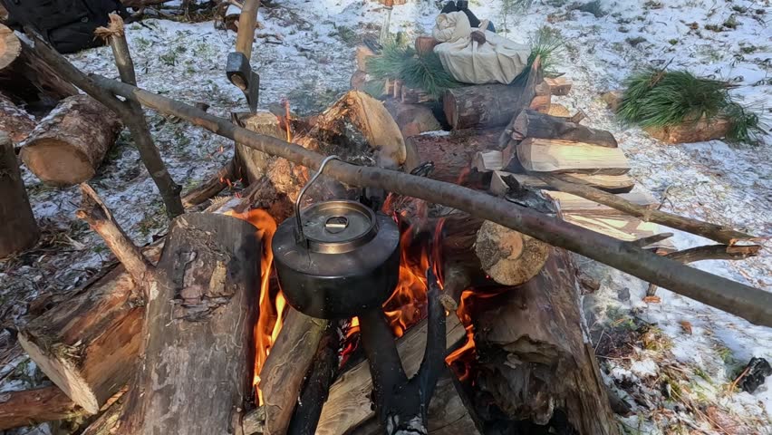 A metal kettle with water is heated on a fire in a winter forest