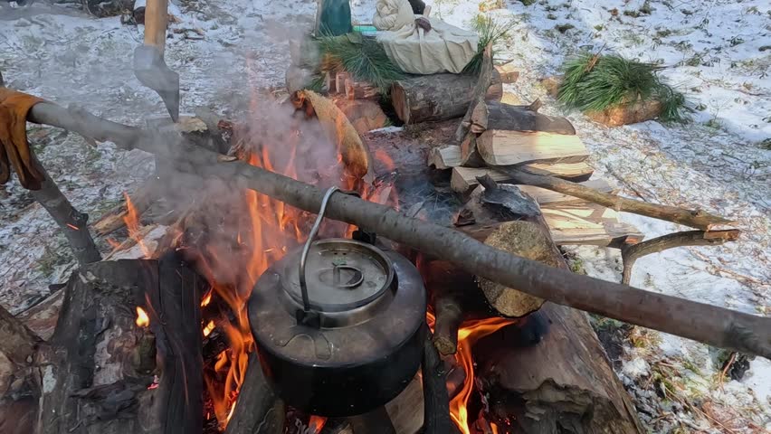 A metal kettle with water is heated on a fire in a winter forest
