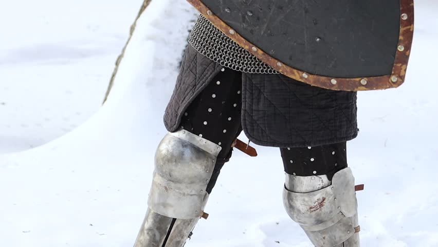 Portrait of a medieval knight in full armor wearing a helmet with a visor and holding a sword and shield in his hands, posing against the backdrop of a winter park.
