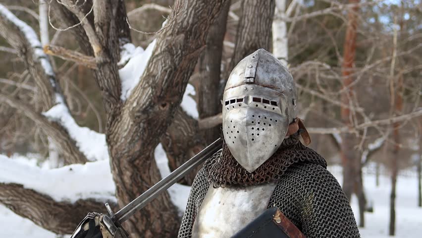 Portrait of a medieval knight in full armor wearing a helmet with a visor and holding a sword and shield in his hands, posing against the backdrop of a winter park.