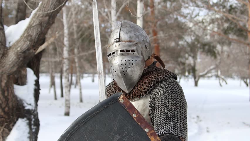 Portrait of a medieval knight in full armor wearing a helmet with a visor and holding a sword and shield in his hands, posing against the backdrop of a winter park.