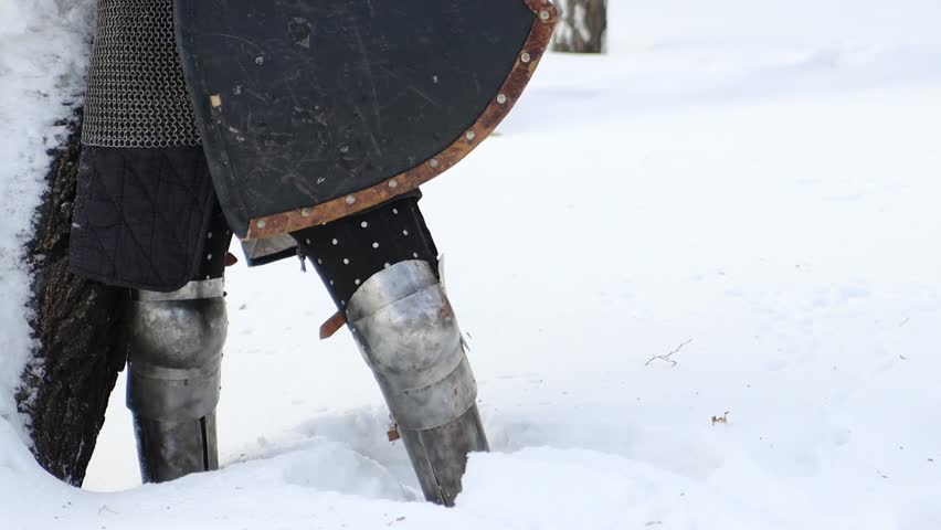 Portrait of a medieval knight in full armor wearing a helmet with a visor and holding a sword and shield in his hands, posing against the backdrop of a winter park.