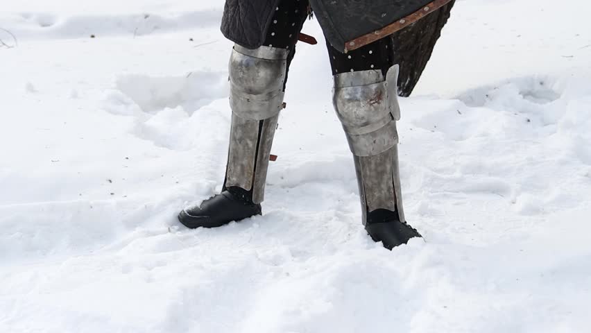 Portrait of a medieval knight in full armor wearing a helmet with a visor and holding a sword and shield in his hands, posing against the backdrop of a winter park.