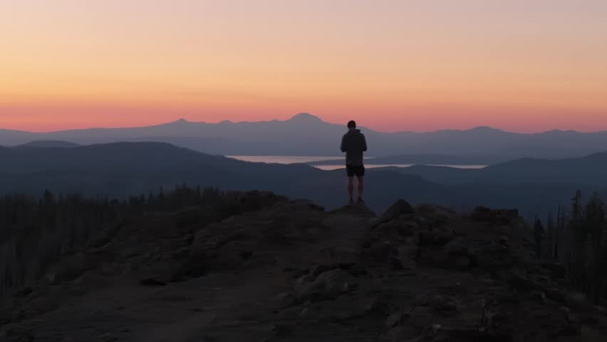 Hiker stands on rocky peak overlooking mountain landscape at colorful sunrise