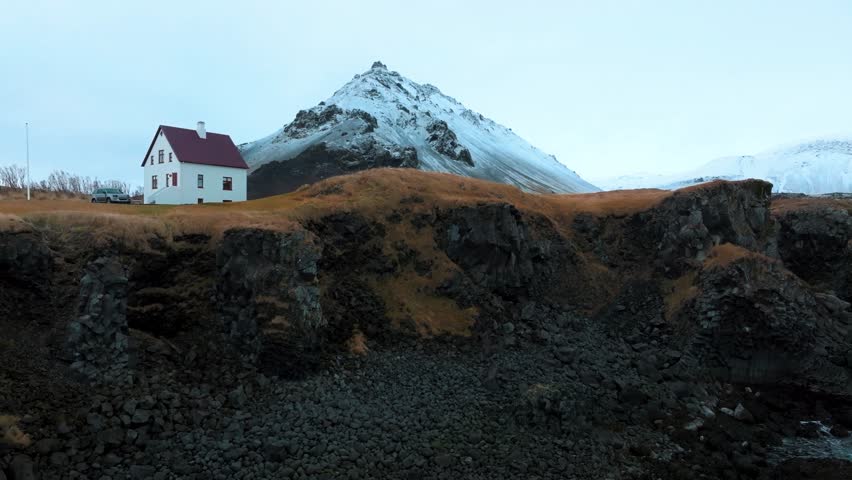 Cozy cottage on a cliff below a snowy cone shaped volcanic mountain in inter in Iceland - ascending aerial reveal