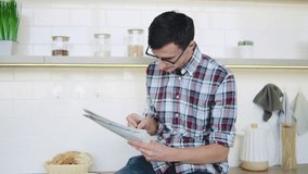 brunette man in a plaid shirt and reading glasses sits in his modern kitchen, reads a newspaper, drinks coffee, and searches for jobs in the classified ads, circling the relevant ones with a pen - Powered by Shutterstock - Get 15% off with code: PIKWIZARD15