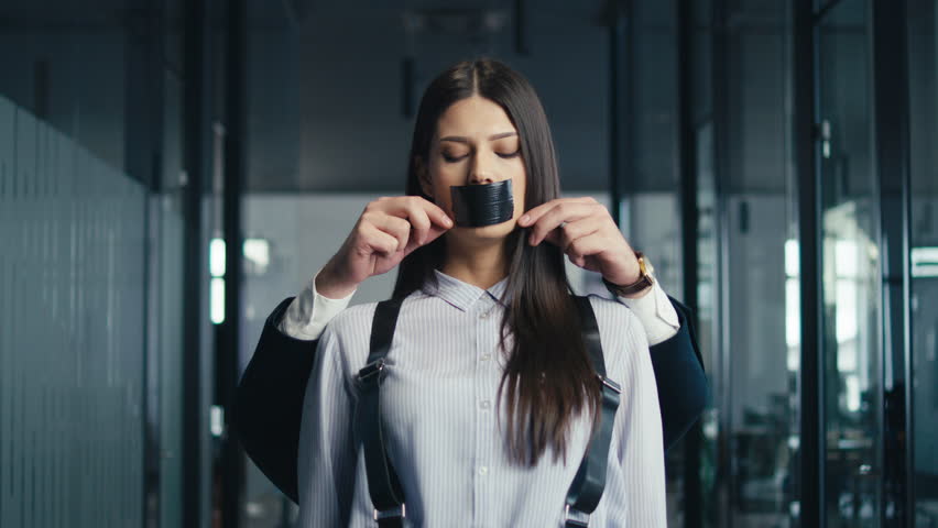 Young professional woman standing in an office corridor with her mouth taped shut by a man