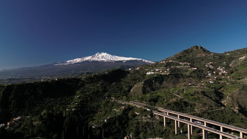 Aerial drone footage of Mount Etna, featuring its snow-capped peak and lush green foothills. A stunning view of Sicily’s iconic volcano, perfect for nature, travel, and documentary projects.