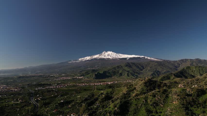 Aerial drone footage of Mount Etna, featuring its snow-capped peak and lush green foothills. A stunning view of Sicily’s iconic volcano, perfect for nature, travel, and documentary projects.