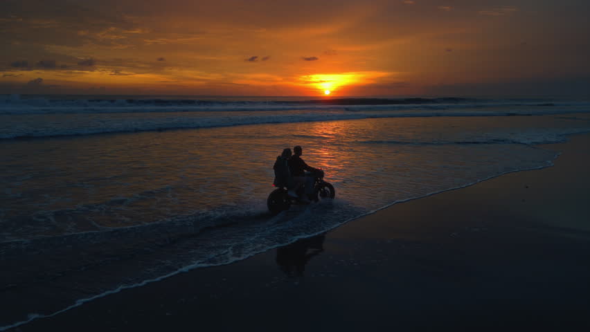 Couple rides retro motorcycle on the beach or shore. Man, woman in love ride a bike in Bali. Trip to ocean or sea at sunset. Travel and road trip. Driving on moped during summer vacation. Aerial drone