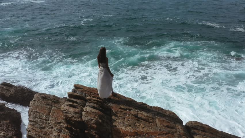 A bride in a flowing white dress stands on a rocky cliff overlooking the ocean in Sri Lanka, with waves crashing below. This high-quality stock photo captures a dramatic and romantic moment, perfect