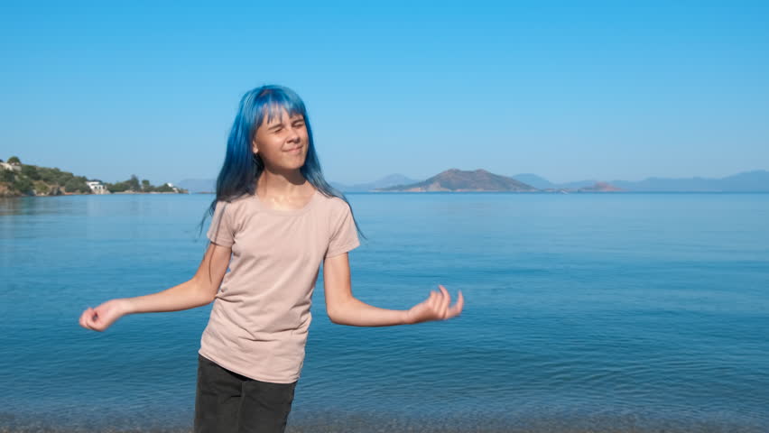 Blue girl makes dancing moves. A happy teen moves on the beach and enjoy the time against blue bay in summer.