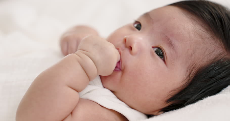 Closeup of newborn baby gently sucking hand on white blanket showing calm expression. Represents self-soothing, comfort, early development, nurturing care, and maternal affection in babyhood.