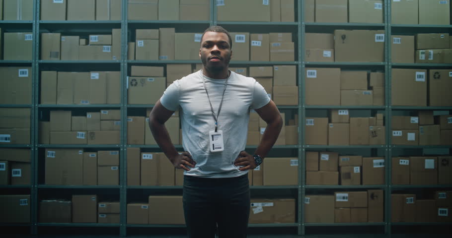 Dolly Shot of Positive African American Warehouse Employee Crossing Arms, Smiling and Looking at Camera. Postal Delivery Service, Logistics Distribution Center with Shelves full of Cardboard Boxes.