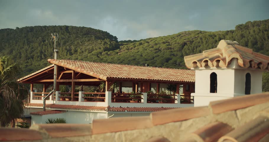 A coastal Mediterranean landscape featuring palm trees, pines, and tiled rooftops against a backdrop of the blue sea and a partly cloudy sky. The view highlights the region