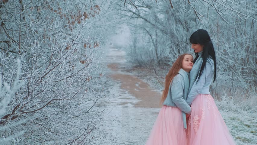 happy family mother and daughter posing for photo shoot, woman hugs little girl with love care, two people together. Winter nature forest trees frost, white snow. Same clothes gray sweater pink dress