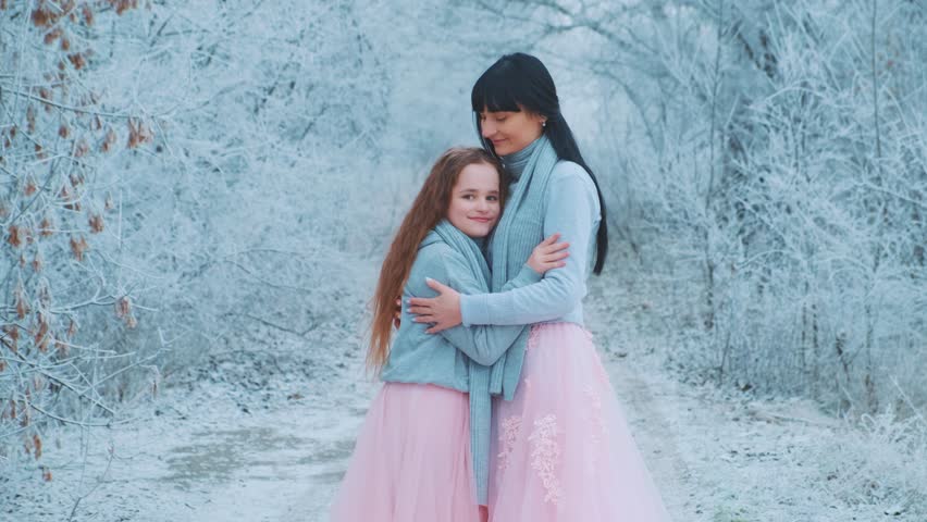 happy family mother and daughter posing for photo shoot, woman hugs little girl with love care, two people together. Winter nature forest trees frost, white snow. Same clothes gray sweater pink dress