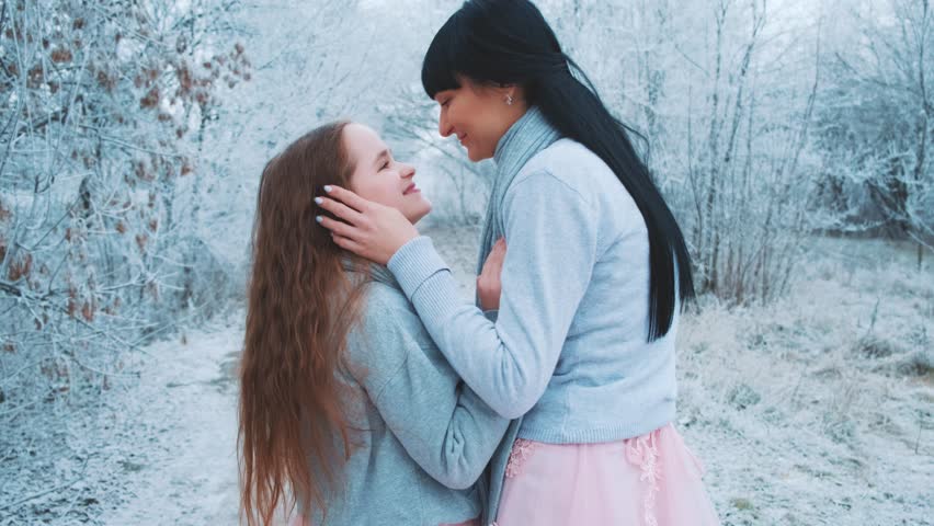 happy family mother and daughter posing kissing hand stroking head hair, woman hugs little girl with love care, two people together. Winter forest trees frost, white snow. Same clothes gray sweater.
