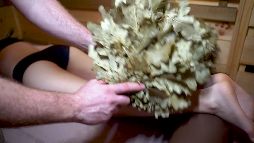 A bearded masseur in a Russian sauna massages a young woman with an oak broom. Relaxing atmosphere in the steam room of the Finnish sauna.