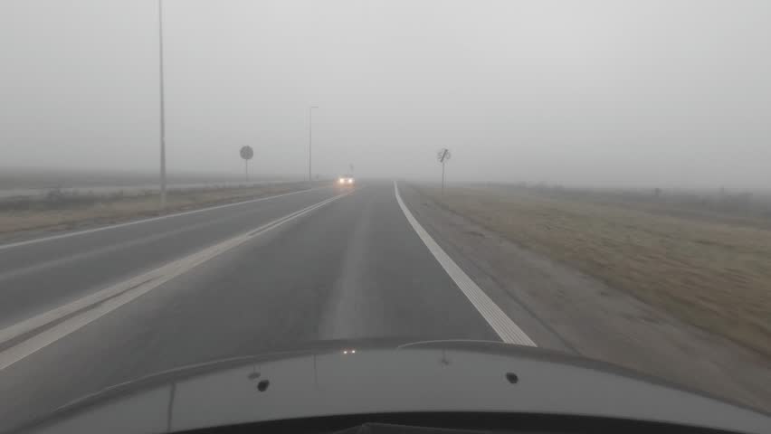 Foggy road view from a car-mounted camera, showing the vehicle, misty asphalt, and passing lit cars. Leafless trees line the autumn roadside.
