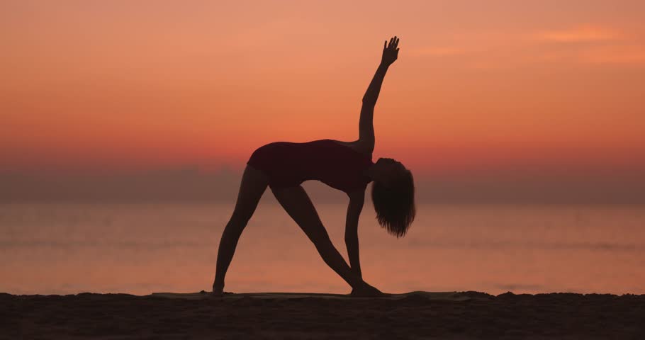 A silhouette of a woman gracefully stretching in a yoga pose on the beach at sunset, with the ocean as the backdrop. Ideal for promoting wellness, mindfulness, flexibility, and outdoor yoga practices.