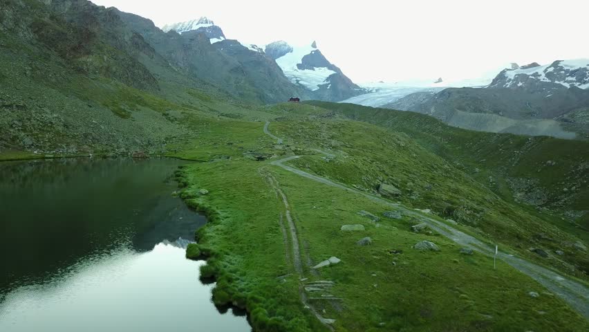 Aerial view of a hiking path winding through the green hillside towards a wooden house, with snowy rocky mountains in the background on a summer morning, in Zermatt, Valais, Switzerland