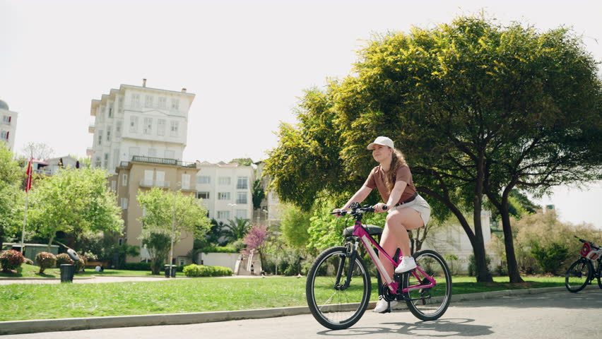 Young woman rides rental bike on Buyukada Island. Female cyclist enjoys scenic cityscape. Casual biking in urban park area. Solo traveler explores historic location on bicycle.