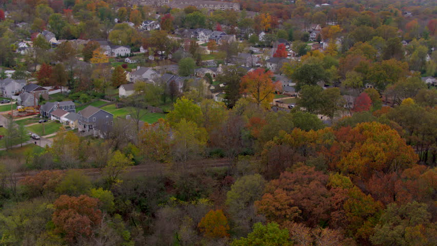 Aerial over train tracks and trees towards houses in Kirkwood neighborhood in St. Louis, Missouri on a cloudy evening in Autumn.