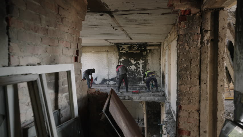 Construction workers dismantle war-damaged apartment, demolish wall with jackhammers. Team cuts reinforcement in debris-filled building for urban renewal. Focus on reconstruction in residential area.