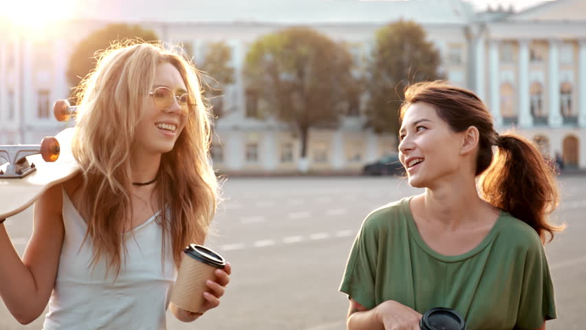 Two fashion hipster girls at outdoors summer party, pretty teenage friends with skateboard in sunshine in street european town laughing and drink coffee. Concept of lifestyle, vintage, friendship