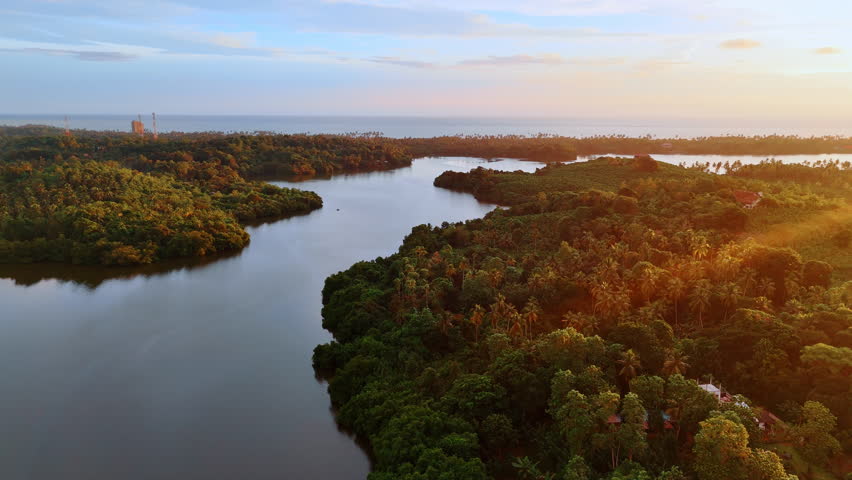Peaceful river flowing among the tropical forests lit with setting sun rays. Waterscape of the Indian Ocean at backdrop. Aerial perspective.