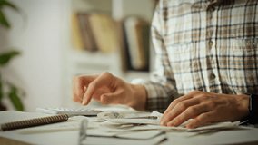A person sorts through various receipts and documents on a cluttered desk, using a calculator and a pen. Plants are visible in the background, creating a cozy workspace atmosphere - Powered by Shutterstock - Get 15% off with code: PIKWIZARD15