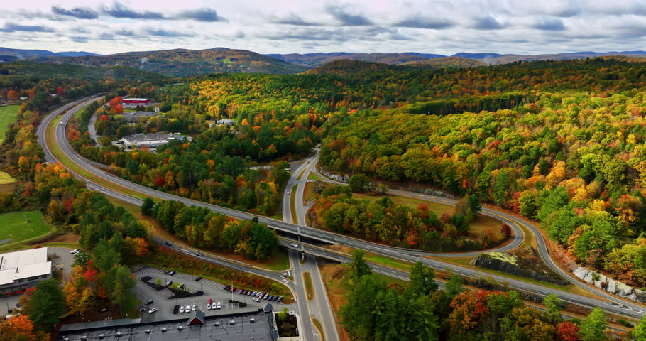 Highways cross the landscapes overgrown with colorful woods. Mountains under the overcast sky at backdrop. Vermont scenery, New England, USA. Aerial view.
