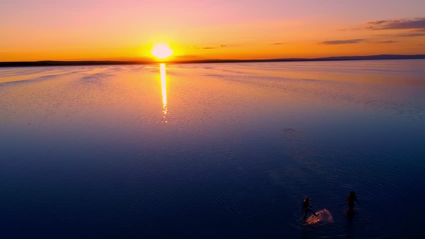 Silhouettes of two people walking on a tranquil lake during a sunset with orange and purple hues. Perfect for nature, serenity, and romance-themed.