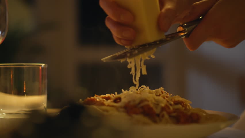 A hand grates cheese over a serving of pasta, adding flavor to the dish. Soft lighting creates a warm atmosphere perfect for an intimate dinner