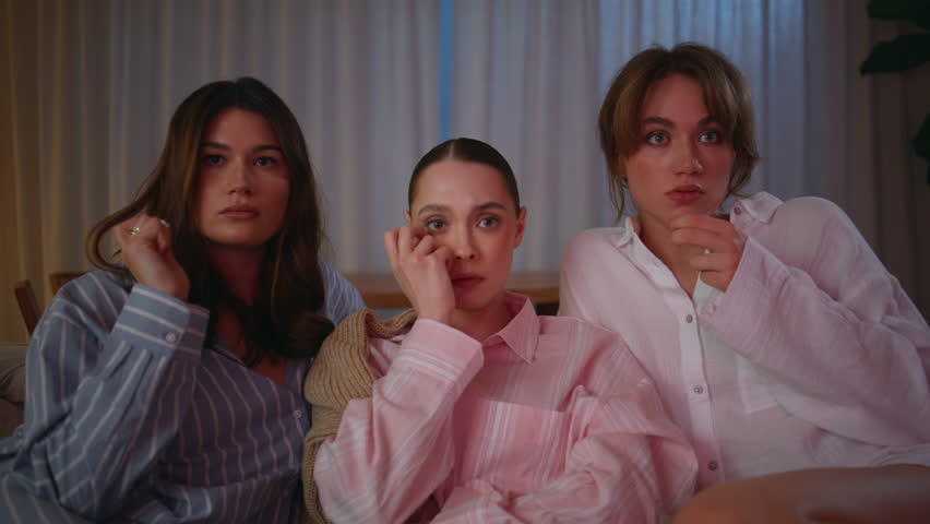 Frightened friends watching movie at night home closeup. Tensed three women scared by horror film eating popcorn in pajamas at evening weekend gathering. Girls reacting to suspenseful moment at couch.