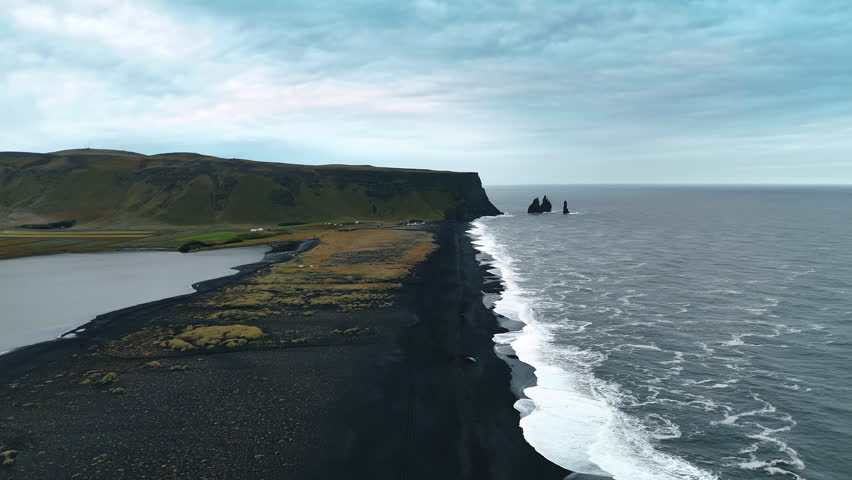 Black narrow sandy beach line stretching to the steep mountain. Few sharp rocks stick out from waterscape. Shore of Iceland on a grey dull day.