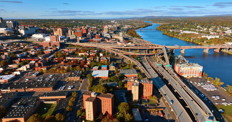 Big interchange with highways, loops and freeways near the waterscape of the river and bridge across it. Sunlit panorama of Albany, New York, USA along the Hudson River. Aerial view.